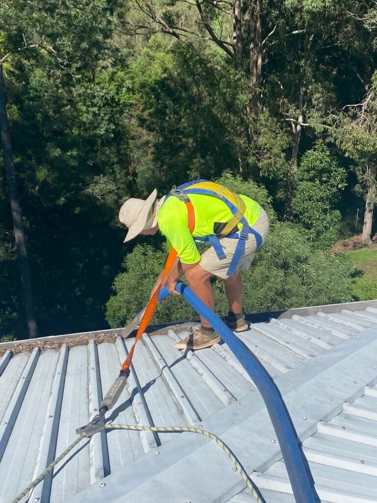 Man on roof cleaning gutters in Brisbane. Wearing hi vis yellow and a hat.